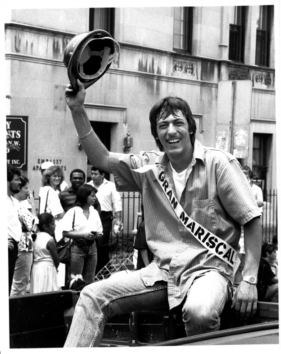 Black and white image of a man waving a construction hat with sash reading “Gran Mariscal” (or “Grand Marshal”) on his chest. 