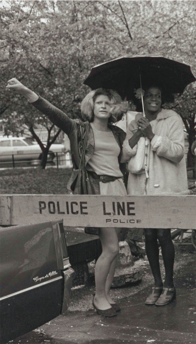 Black and white photo of Sylvia Rivera, fist raised, and Marsha P. Johnson, holding umbrella, in front of wooden police barricade.