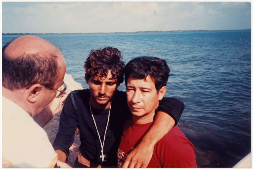 Photo of three men, arms across each other's sholders, standing on patch of land in vast blue ocean