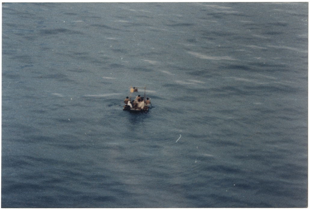 Birds-eye blurry photo of small raft carrying perhaps five people on vast blue ocean