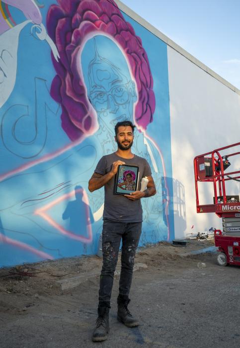 Foto a color del muralista Albert "Tino" Ortega de pie frente al mural esbozado para Jailah Nicole Silguero