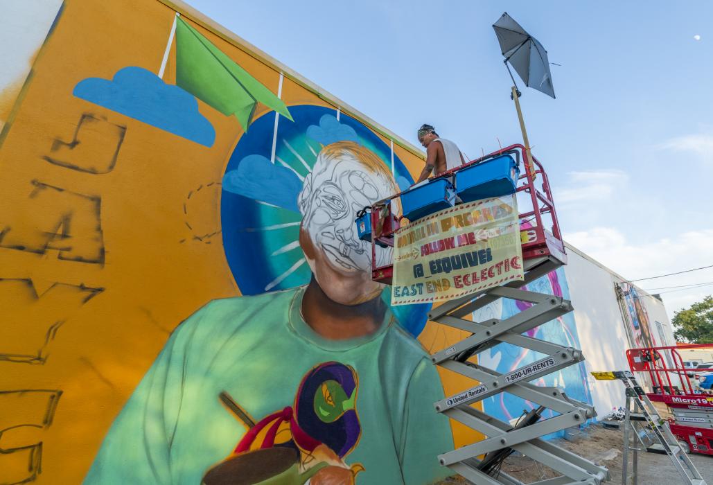 Foto a color del muralista Ruben Esquivel en un elevador de tijera frente al retrato mural de Jayce Luevanos