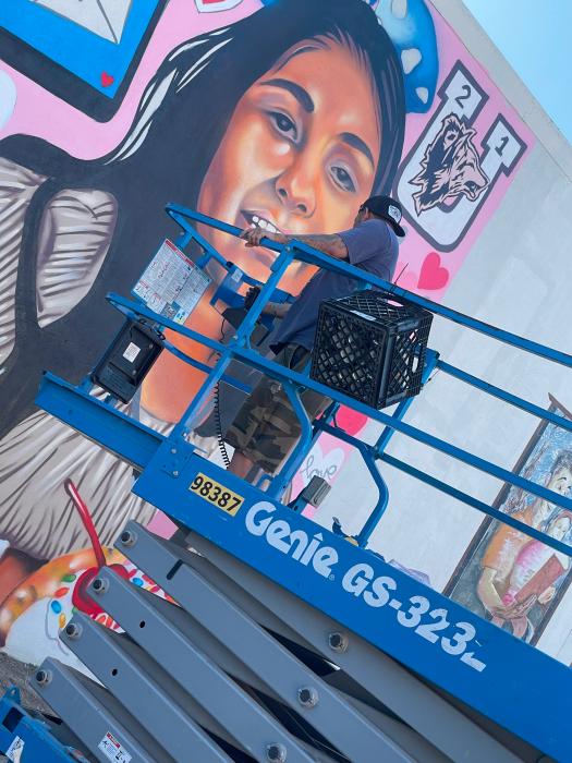 Foto a color del muralista Joey C. Martinez en un elevador de tijera azul frente al retrato mural de Annabell Rodriguez
