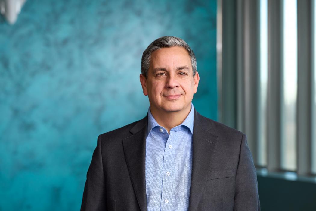 Professional portrait of a smiling individual in a business suit, standing in front of a turquoise textured background.