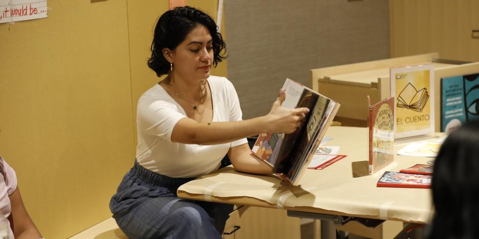 Women with short dark hair is seating down in a bench while reading a book to an audience