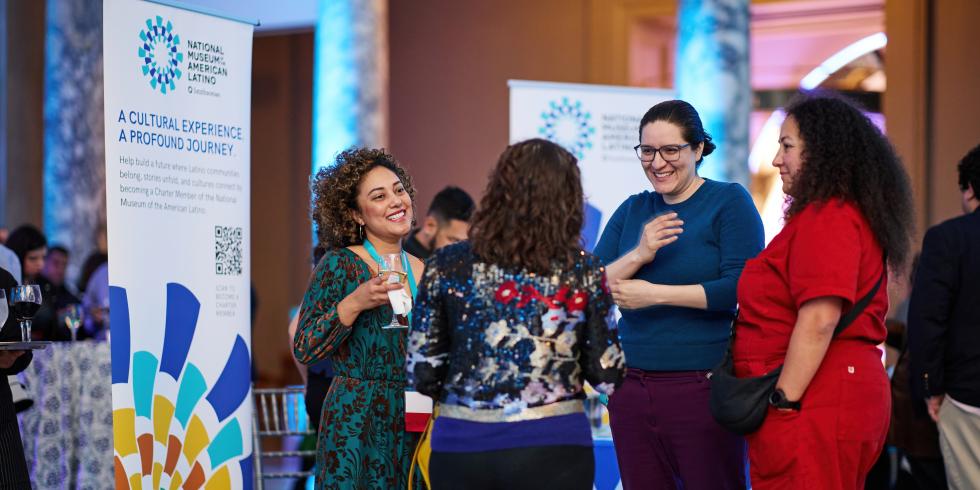 Four women standing in a circle having a conversation in a large room. The two women facing the camera have smiles on their faces. To the side of the group is a vertical banner containing the National Museum of the American Latino logo..