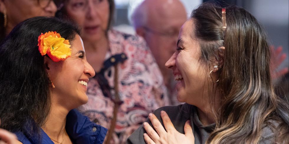 Two women facing each other with big smiles on their face. Woman on left has long black hair with a yellow, orange, and red flower tucked behind her ear wearing a blue dress. Women on right has long brunette hair and a gray blouse. There are people standing behind and in front of the women.