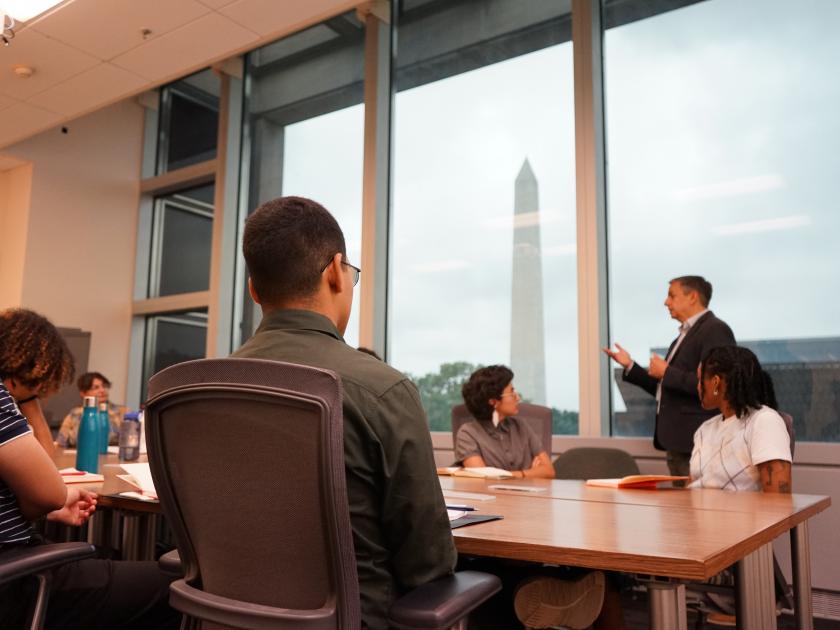 Several interns are seated in a conference room. Jorge Zamanillo, Founding Director of the National Museum of the American Latino, speaks to the interns. 