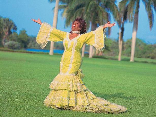 Celia Cruz wearing a yellow rumba dress with ruffles and wide sleeves. Her arms are outstretched. Behind her there are palm trees.