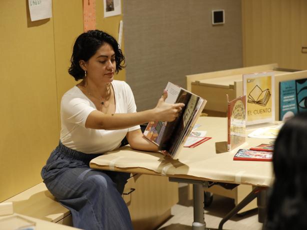 Women with short dark hair is seating down in a bench while reading a book to an audience