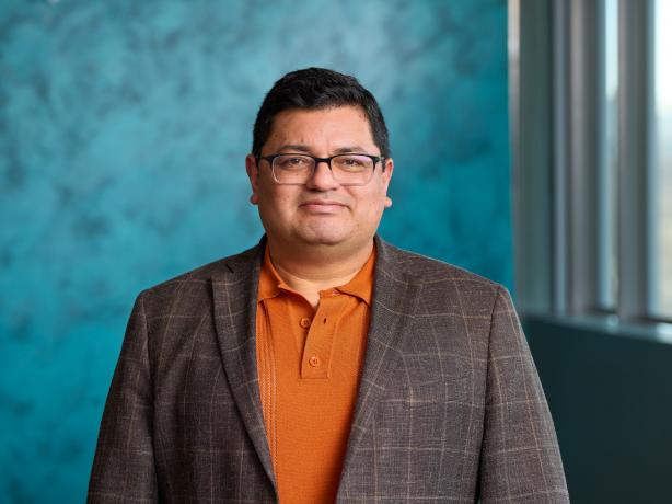Person wearing glasses, an orange shirt, and a patterned blazer, smiling and standing in front of a blue textured background.