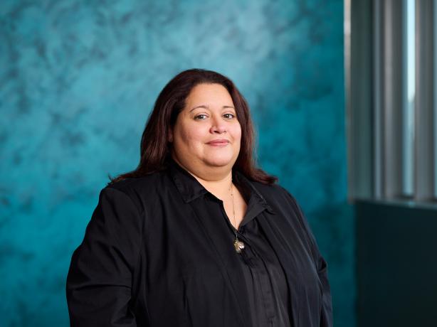 Professional portrait of an individual wearing a black shirt, standing in an office environment with a textured blue background. The person exhibits a subtle smile and has shoulder-length dark hair.