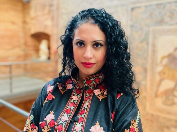 A person wearing a traditional floral embroidered coat standing in front of a mosaic-tiled wall inside a historic building.