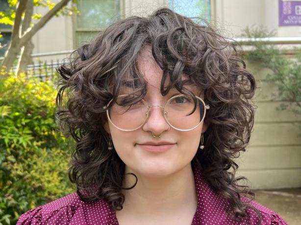 A person with curly hair wearing glasses standing outside.