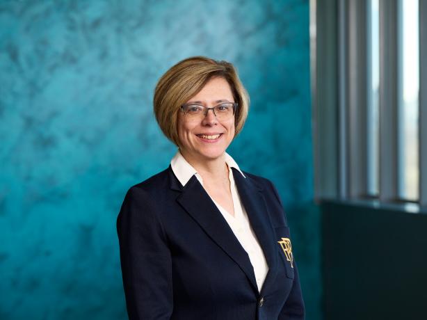 A professional portrait of an individual wearing a dark blazer and a white shirt, smiling against a textured turquoise background near a window.