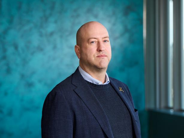 A professional portrait of an individual standing confidently against a teal backdrop, wearing a dark blazer over a buttoned shirt.