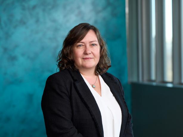 Professional portrait of a person standing indoors against a textured turquoise wall. The individual is wearing a dark blazer over a white top, and a turquoise necklace. Natural light from the right side subtly emphasizes the facial features.