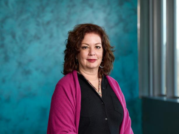 A professional portrait of a person wearing a pink cardigan over a black outfit, standing in front of a textured turquoise background.