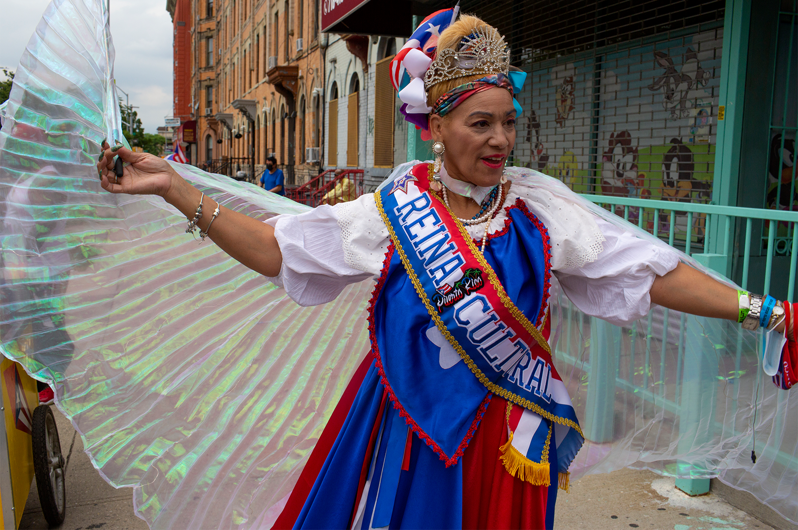 00625-D2-2021.09.29 Color photo of a woman in a parade with extended arms holding pleated, iridescent wings with halter neck.
