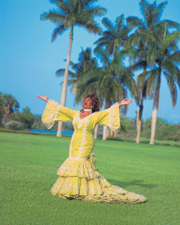 Celia Cruz Celia Cruz wearing a yellow rumba dress with ruffles and wide sleeves. Her arms are outstretched. Behind her there are palm trees.