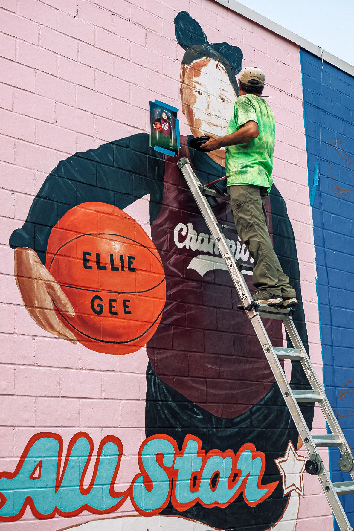 L2-0001145-D1-2022.10.10 Color photo of muralist Abel Ortiz on a ladder finishing up details of portrait mural for Eliahna "Ellie" Amyah Garcia.