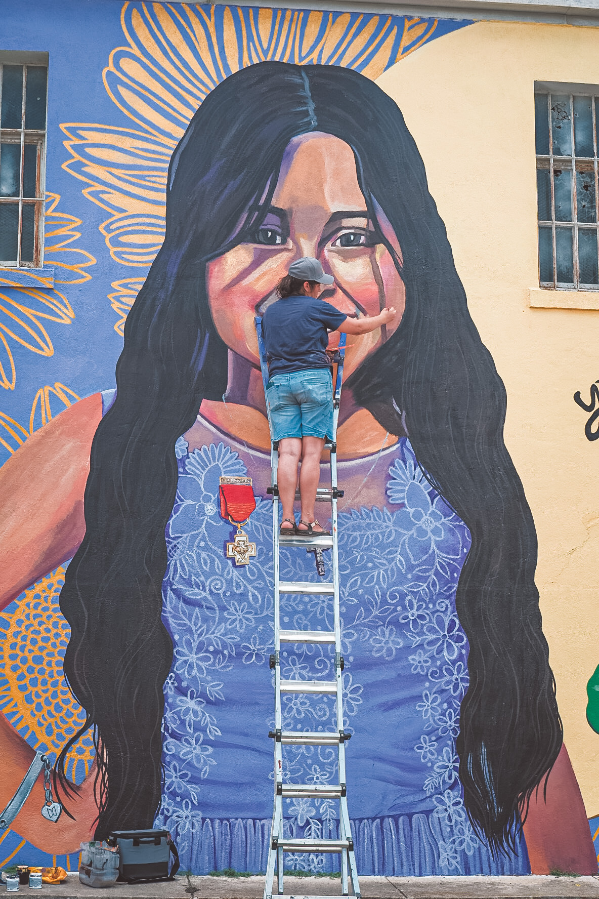 L2-0001146-D1-2022.10.10 Foto a color de la muralista Cristina Noriega en una escalera agregando los toques finales del retrato mural de Amerie Jo Garza
