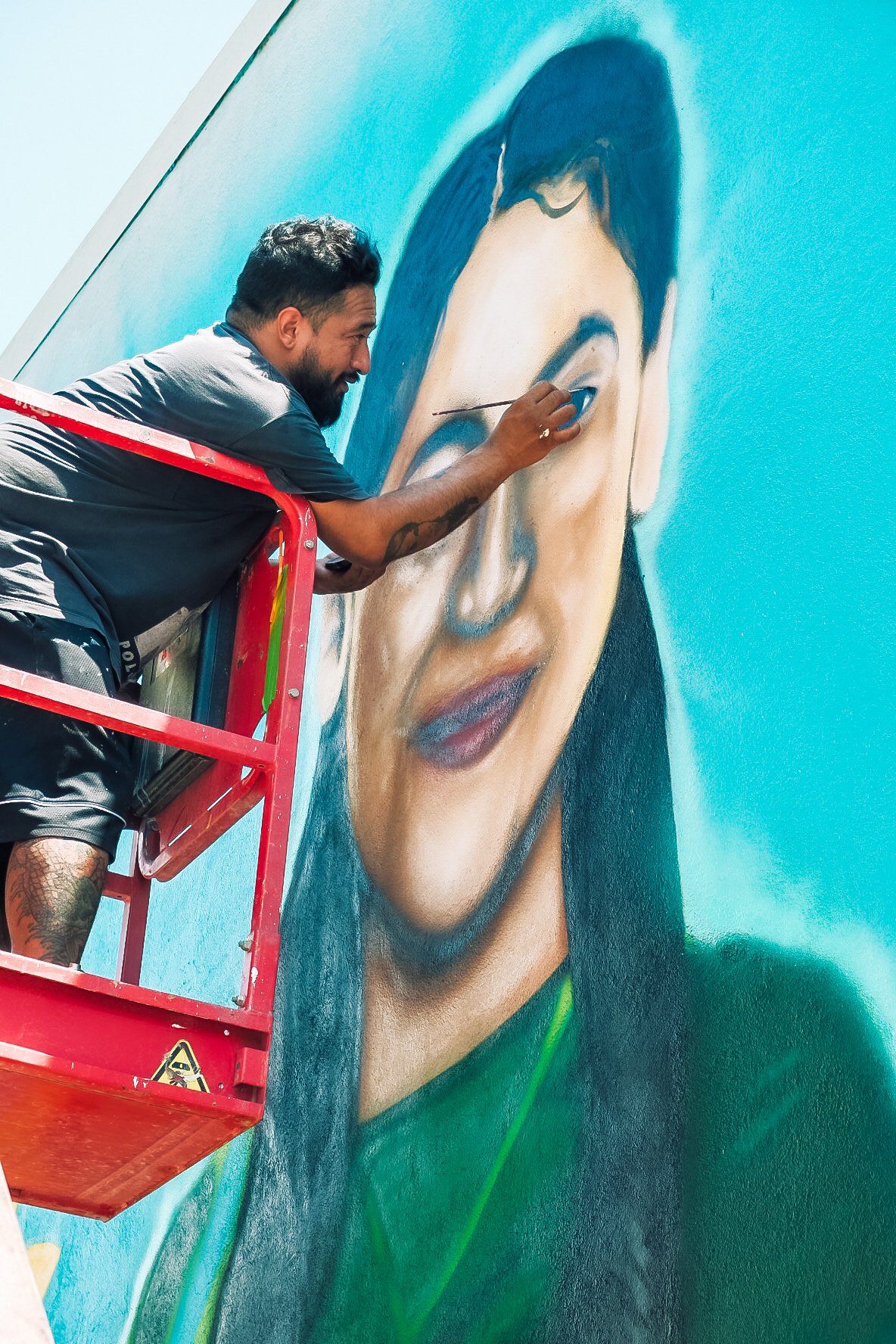 L2-0001148-D1-2022.10.10 Color photo of muralist Filiberto Mendieta on a scissor lift painting the eyes on portrait mural for Eliahna Torres.