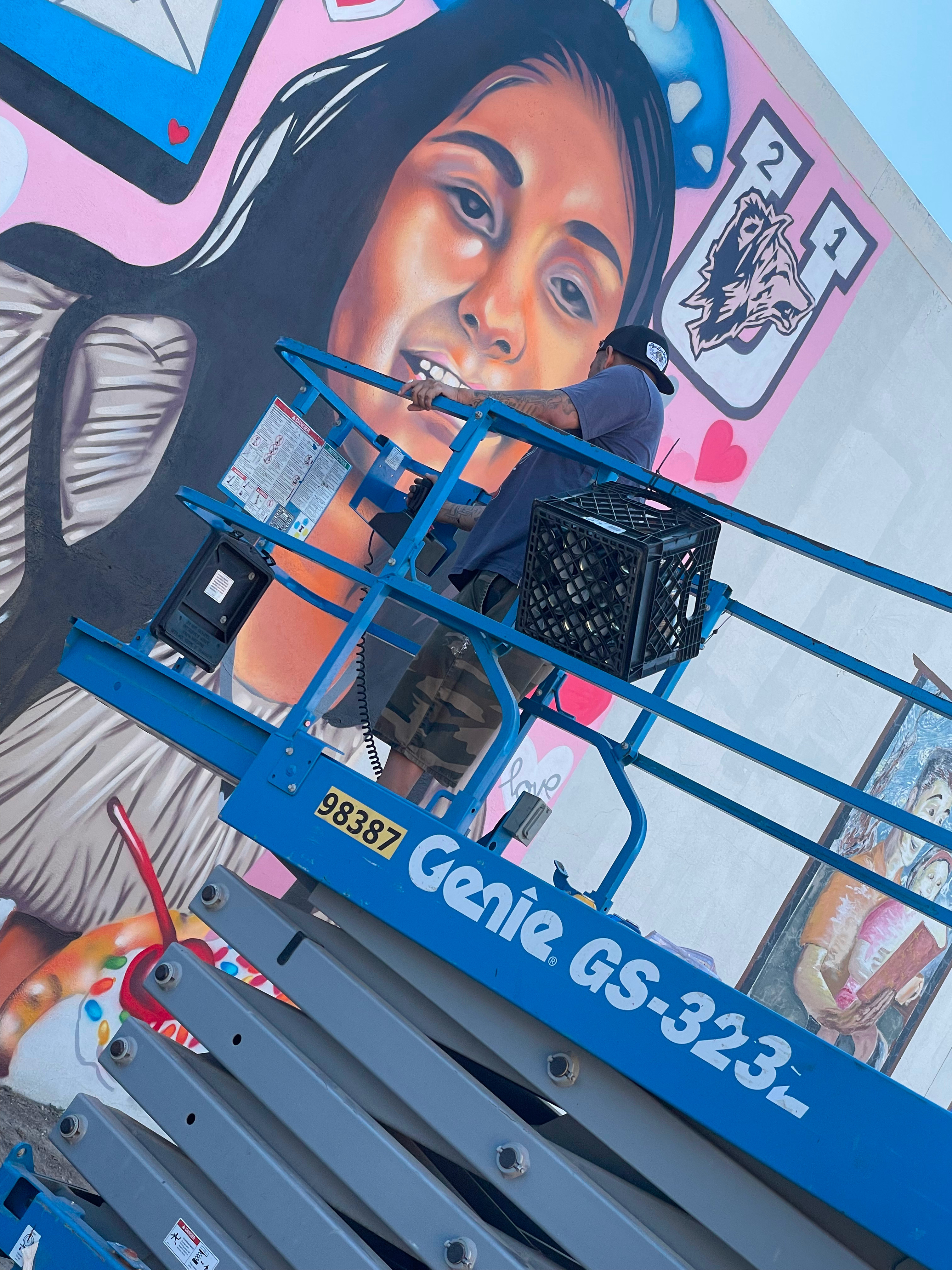 L2-0001163-D1-2022.10.13 Color photo of muralist Joey C. Martinez on a blue scissor lift in front of portrait mural for Annabell Rodriguez.