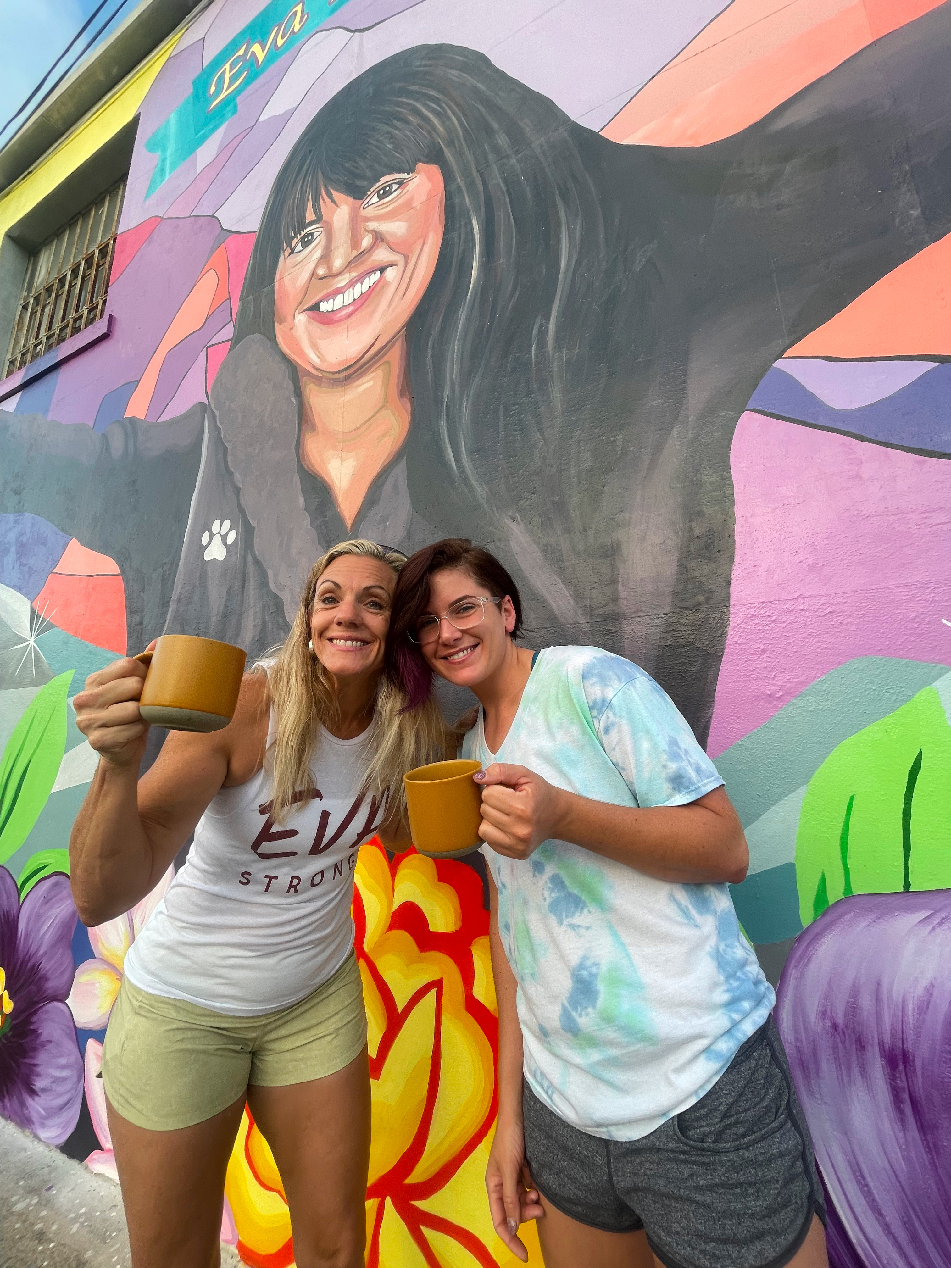 L2-0001164-D1-2022.10.13 Color photo of two people standng in front of Eva Mireles' portrait mural holding a mug in their hands.