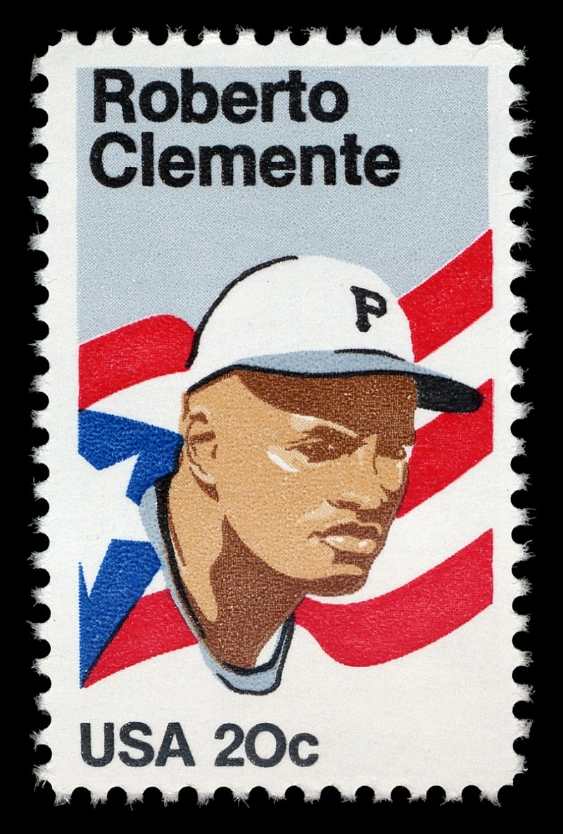 Roberto Clemente USPS stamp of baseball player Roberto Clemente. The stamp displays an illustration of the face of Clemente wearing a baseball hat with a P on it, emblematic of the Pittsburgh Pirates. In the background, there is a Puerto Rican Flag.